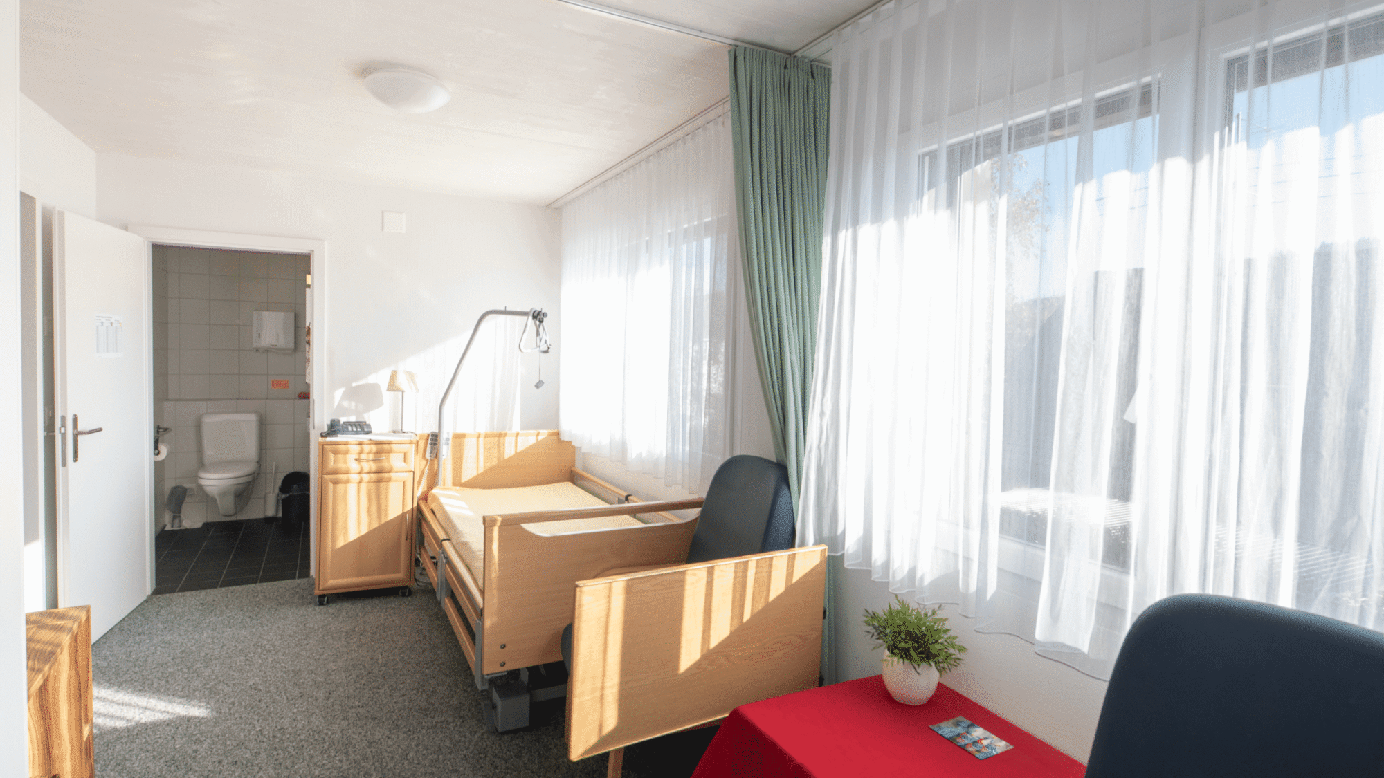 Helles Einzelzimmer mit Pflegebett, Tisch, Stuhl und eigenem Bad.